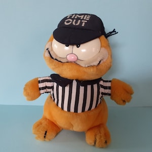 Könnte beinhalten: Ein Plüschtier Garfield, das ein schwarz-weiß gestreiftes Schiedsrichtertrikot und einen schwarzen Hut mit der Aufschrift "Time Out" in weißen Buchstaben trägt.