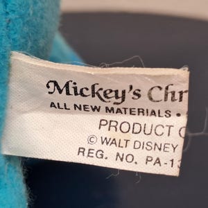 May include: A close-up of a white tag with the text "Mickey's Chr" and "ALL NEW MATERIALS." The tag also includes "PRODUCT O" and "&copy; WALT DISNEY REG. NO. PA-13" on a blue plush toy.
