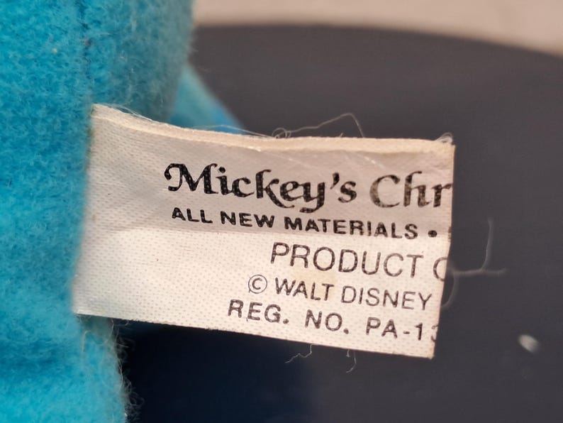 May include: A close-up of a white tag with the text "Mickey's Chr" and "ALL NEW MATERIALS." The tag also includes "PRODUCT O" and "&copy; WALT DISNEY REG. NO. PA-13" on a blue plush toy.