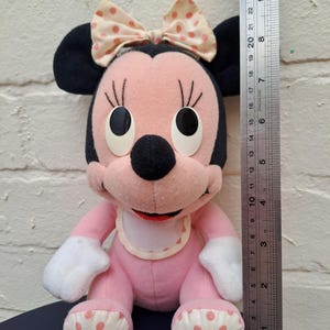 May include: A vintage Minnie Mouse plush toy, primarily pink with black ears and a bow. The toy has white gloves and polka-dot accents on the feet. The plush is approximately 10 inches tall, as measured by a ruler.