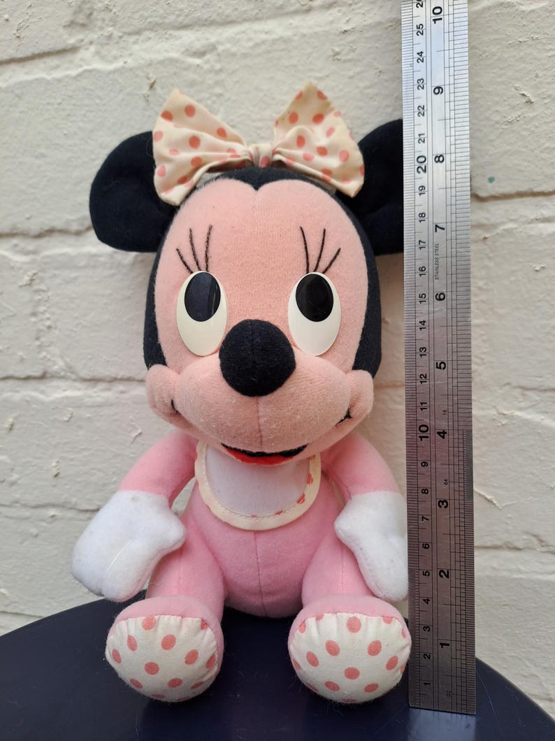 May include: A vintage Minnie Mouse plush toy, primarily pink with black ears and a bow. The toy has white gloves and polka-dot accents on the feet. The plush is approximately 10 inches tall, as measured by a ruler.