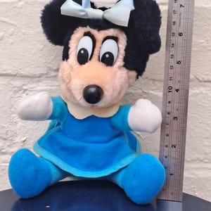 May include: A plush Minnie Mouse doll, featuring black ears, a light blue bow, and a blue dress. The doll has a pink face, white gloves, and blue shoes. A ruler is next to the doll.