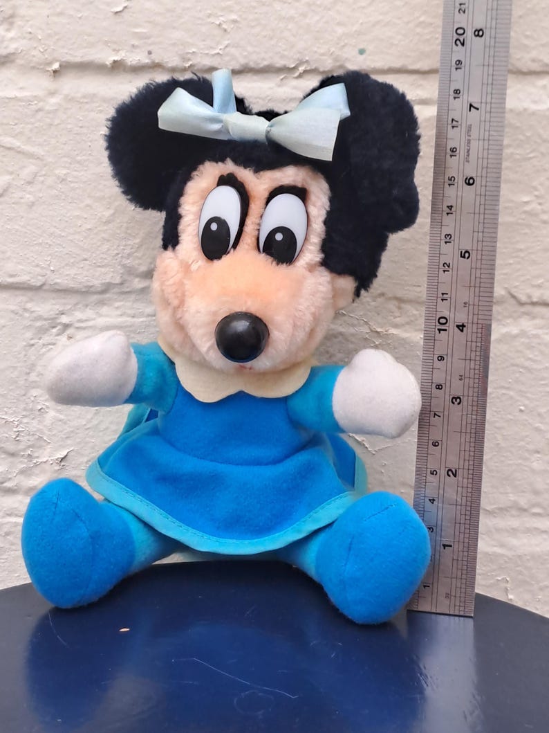 May include: A plush Minnie Mouse doll, featuring black ears, a light blue bow, and a blue dress. The doll has a pink face, white gloves, and blue shoes. A ruler is next to the doll.