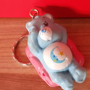May include: Blue Care Bear keychain with a pink sleeping bag. The bear has a crescent moon and star on its belly and is holding its paws up to its face.