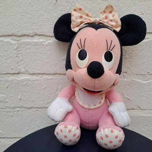 May include: A plush Minnie Mouse doll, featuring a pink body, black ears, and a bow with pink polka dots. The doll has white gloves and polka-dotted feet. It is sitting on a dark blue surface.