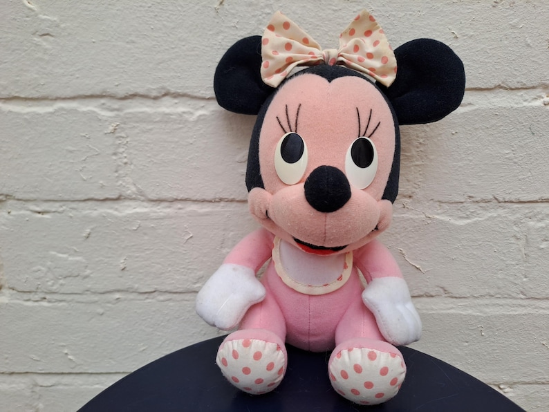 May include: A plush Minnie Mouse doll, featuring a pink body, black ears, and a bow with pink polka dots. The doll has white gloves and polka-dotted feet. It is sitting on a dark blue surface.