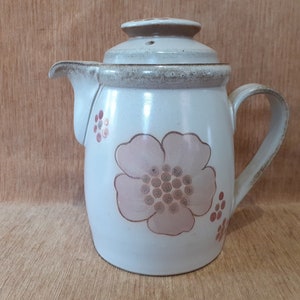 Puede incluir: Una tetera de cerámica blanca con una tapa marrón y un diseño floral. La tetera tiene un asa y un pico. El diseño floral presenta flores rosas con centros marrones y puntos marrones.