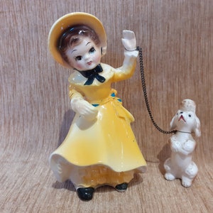 Puede incluir: Una figurita de porcelana de una niña con un vestido amarillo y un sombrero, sosteniendo una correa unida a una figurita de perro de porcelana blanca.