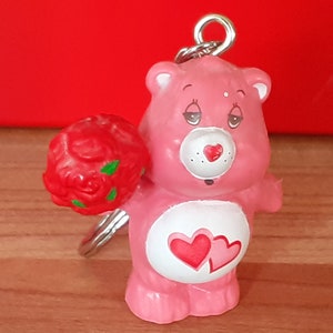 May include: Pink Care Bear keychain with a red rose bouquet and two hearts on its belly.