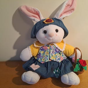 Op de afbeelding: Een witte pluche konijn met een blauwe denim hoed, een geel shirt met een bloemenpatroon en een blauwe denim rok met patchwork. Het konijn houdt een bruine mand met een rode roos.