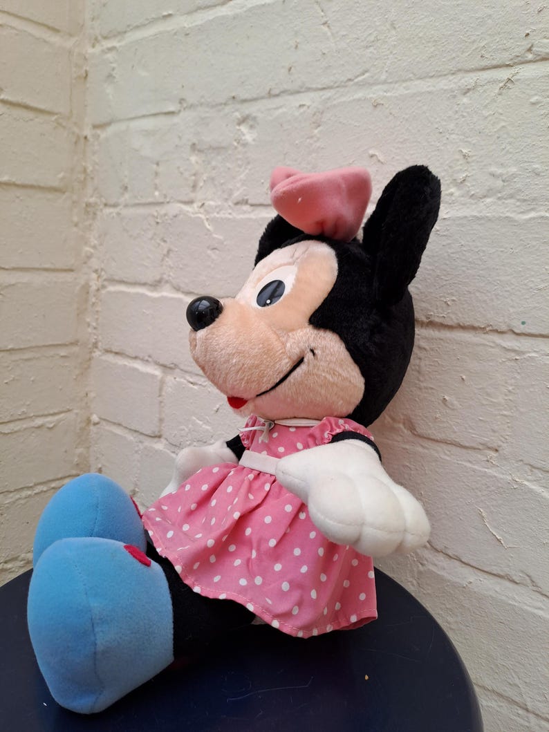 May include: A plush Minnie Mouse doll, featuring black ears, a pink bow, and a pink dress with white polka dots. The doll has white gloves, blue feet, and a smiling expression. A classic Disney character.