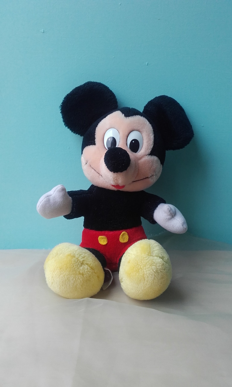 May include: A plush Mickey Mouse toy with black fur, large ears, and a red and yellow outfit. The toy is sitting on a white surface.