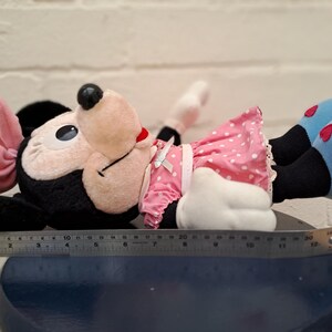 May include: A plush Minnie Mouse doll, lying down, features a black head and ears, a pink bow, and a pink polka-dot dress. Her blue shoes have red heart accents. The doll is approximately 10 inches long.
