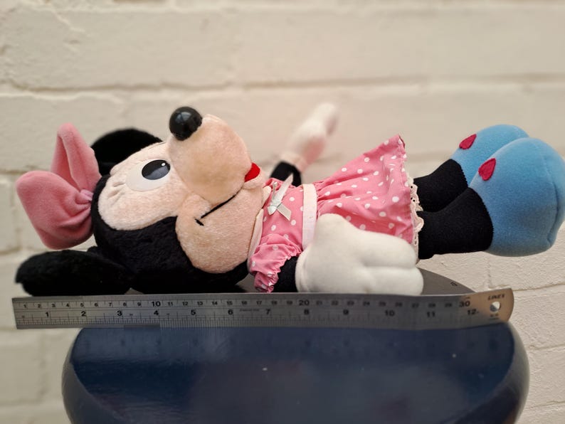 May include: A plush Minnie Mouse doll, lying down, features a black head and ears, a pink bow, and a pink polka-dot dress. Her blue shoes have red heart accents. The doll is approximately 10 inches long.