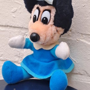 May include: A plush doll of a cartoon mouse character. The doll has a black head with a light blue bow, large eyes, and a black nose. It wears a blue dress and shoes with white gloves.
