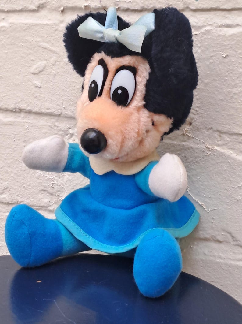 May include: A plush doll of a cartoon mouse character. The doll has a black head with a light blue bow, large eyes, and a black nose. It wears a blue dress and shoes with white gloves.