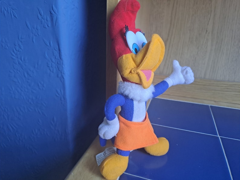 May include: A plush toy of Woody Woodpecker, a cartoon character. The toy is wearing a blue and white shirt, orange shorts, and a white fur collar. The toy has a red head, yellow beak, and blue eyes.