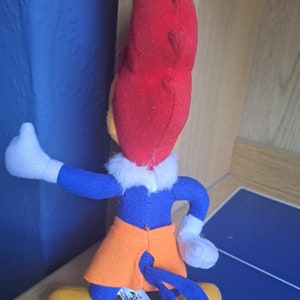 May include: A plush toy of Woody Woodpecker, a cartoon character. The toy is red, blue, and orange. The toy is wearing a blue shirt, orange shorts, and has a red head with a yellow beak. The toy is standing on a blue and white tiled floor.