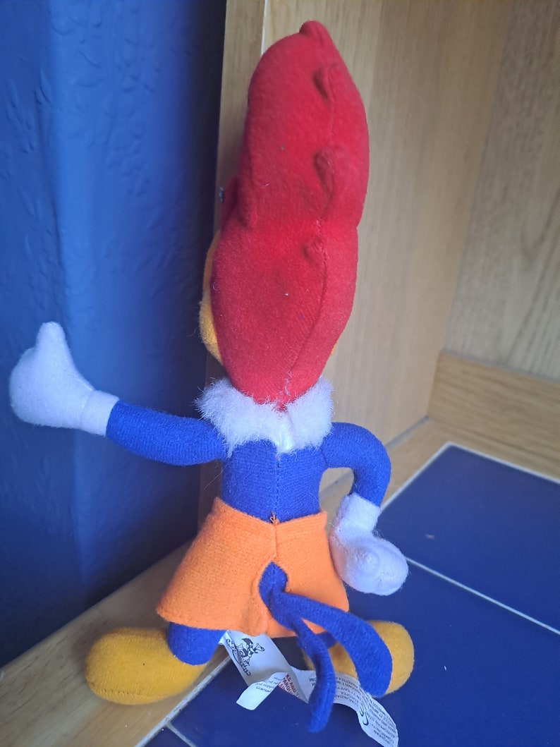May include: A plush toy of Woody Woodpecker, a cartoon character. The toy is red, blue, and orange. The toy is wearing a blue shirt, orange shorts, and has a red head with a yellow beak. The toy is standing on a blue and white tiled floor.