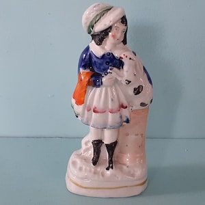 Puede incluir: Una figurita de porcelana de una niña con un vestido azul y blanco que sostiene un perro blanco. La niña lleva un sombrero blanco con detalles verdes y rojos. La figurita está de pie sobre una base blanca con un borde dorado.