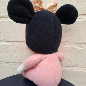 May include: A plush doll of a cartoon character, featuring large black ears and a pink body. The doll has a bow with pink polka dots. The doll is sitting on a dark blue surface.