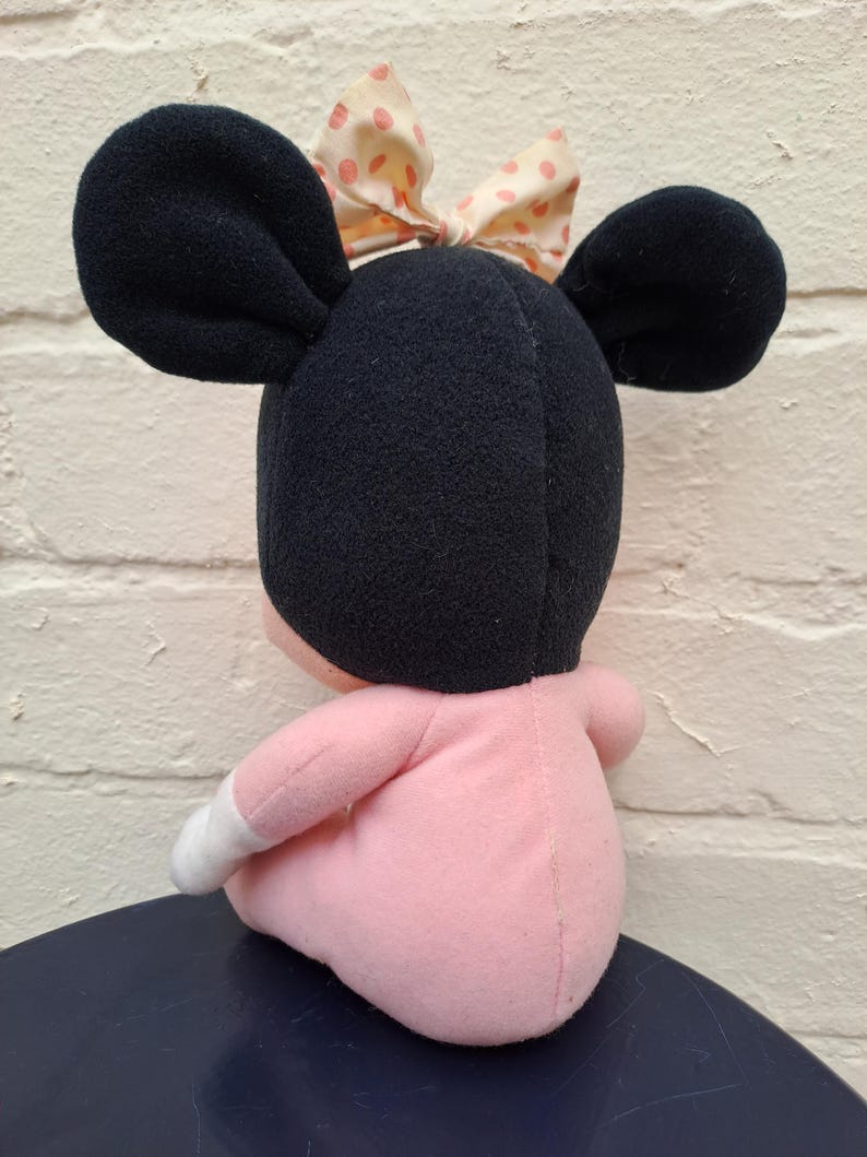 May include: A plush doll of a cartoon character, featuring large black ears and a pink body. The doll has a bow with pink polka dots. The doll is sitting on a dark blue surface.