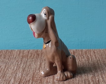 May include: A ceramic figurine of a brown and white bloodhound dog with a black collar and a yellow tag. The dog is sitting with its head tilted to the side and its tongue sticking out.