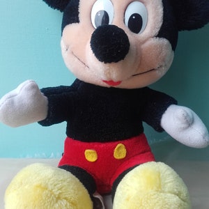 May include: A plush Mickey Mouse toy with black fur, large ears, and a red and yellow outfit. The toy has a friendly expression with black eyes and a red mouth.