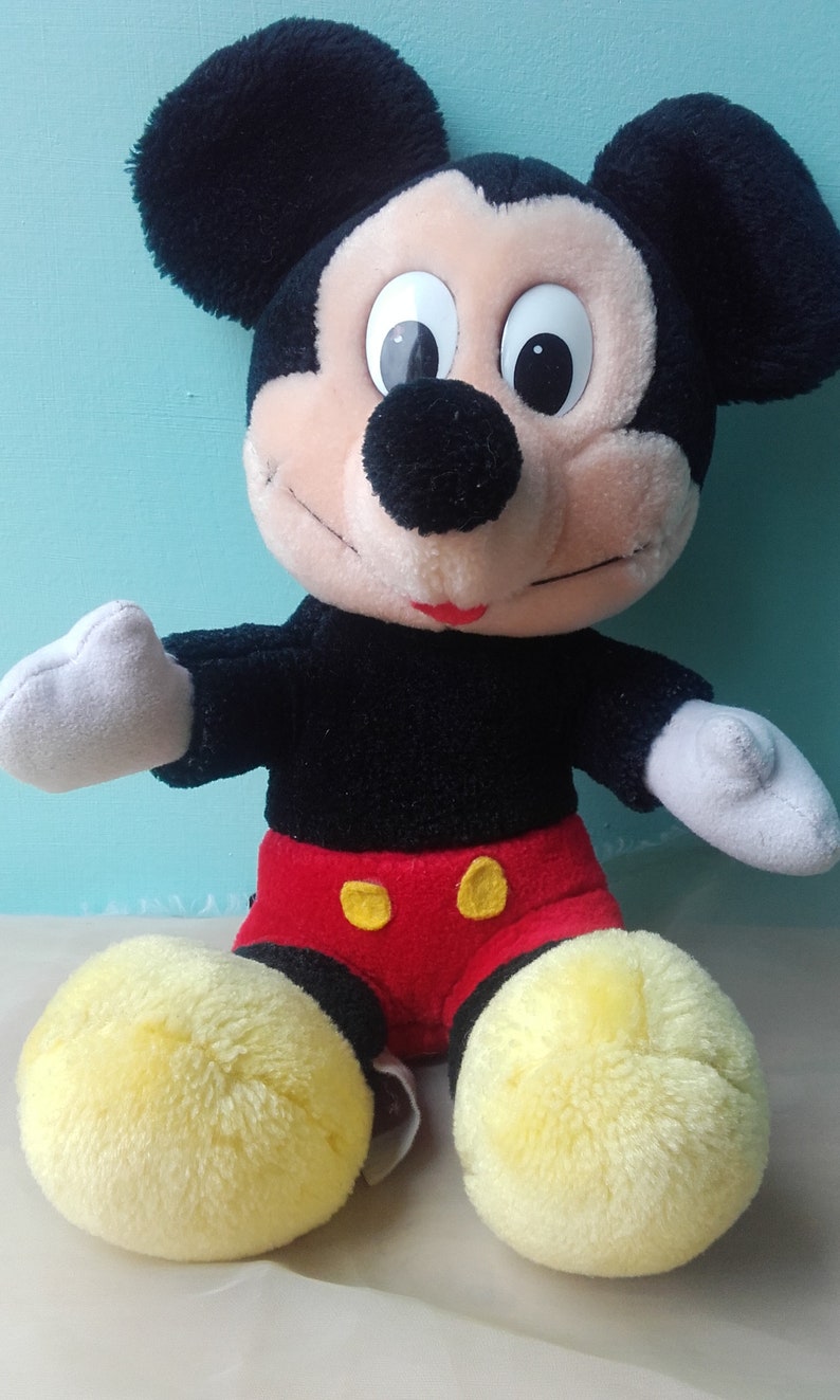 May include: A plush Mickey Mouse toy with black fur, large ears, and a red and yellow outfit. The toy has a friendly expression with black eyes and a red mouth.