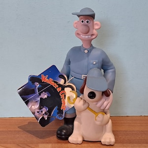 Puede incluir: Un juego de figuras de Wallace y Gromit. Wallace viste un uniforme y gorra azules, mientras que Gromit sostiene una llave inglesa amarilla. Una etiqueta con el logotipo de Wallace & Gromit está adjunta. Las figuras están sobre un fondo azul claro.