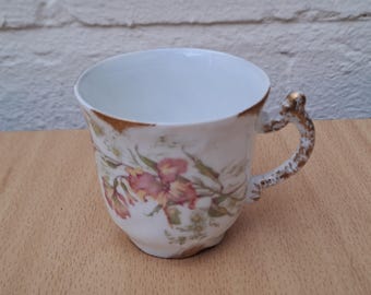 May include: White porcelain teacup with a floral design in pink and green. The cup has a gold-colored rim and handle, and sits on a wooden surface. The teacup is likely vintage or antique.