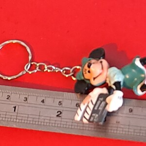 May include: A Mickey Mouse keychain figurine in a playful pose, featuring a silver keyring and chain. The figure is primarily green, black, and white, with a smiling face. The keychain is approximately 2 inches long.