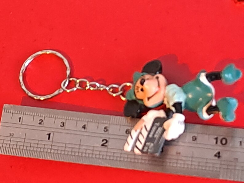 May include: A Mickey Mouse keychain figurine in a playful pose, featuring a silver keyring and chain. The figure is primarily green, black, and white, with a smiling face. The keychain is approximately 2 inches long.
