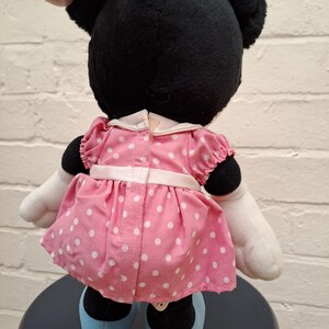 May include: A plush Minnie Mouse doll, viewed from the back, with black fur, large ears, and a pink bow. She wears a pink dress with white polka dots, white gloves, and blue shoes. The doll is sitting on a dark blue stool.