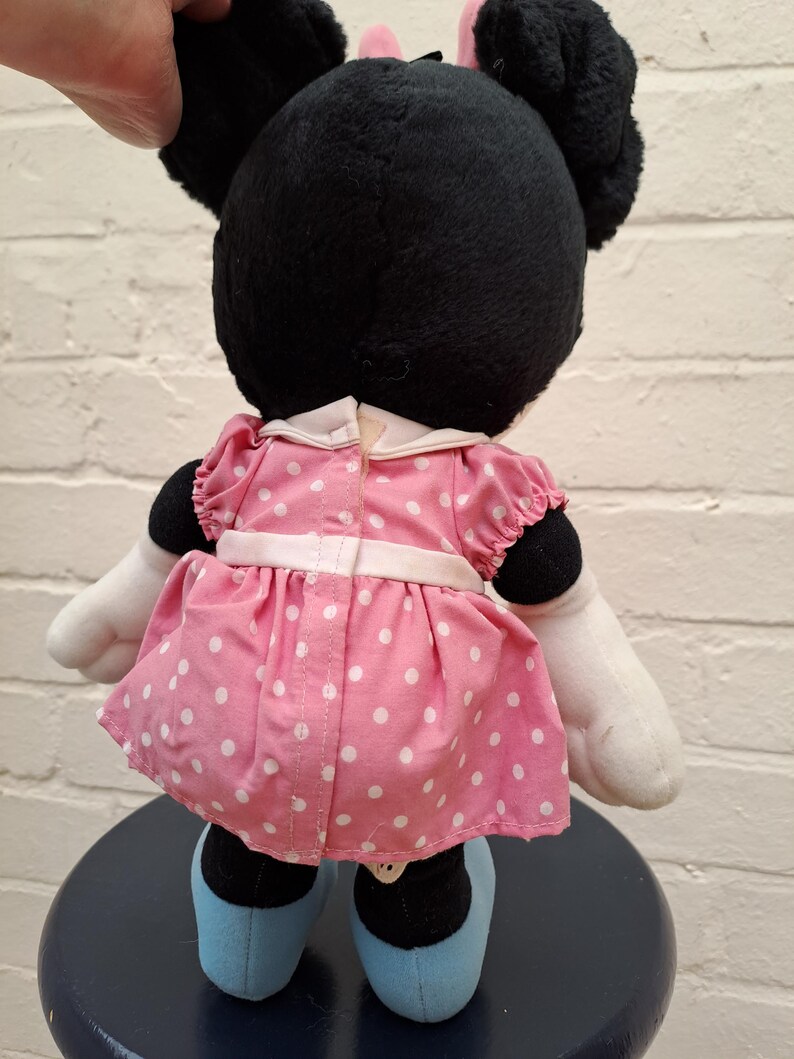 May include: A plush Minnie Mouse doll, viewed from the back, with black fur, large ears, and a pink bow. She wears a pink dress with white polka dots, white gloves, and blue shoes. The doll is sitting on a dark blue stool.