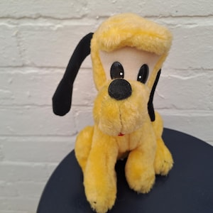 Puede incluir: Un peluche vintage de Pluto. El perro de peluche tiene pelaje amarillo, orejas negras y una nariz negra. Los ojos son grandes y negros. El juguete está sentado sobre una superficie azul oscuro contra una pared de ladrillos blancos.