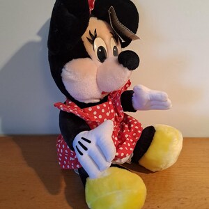 May include: A plush Minnie Mouse toy wearing a red dress with white polka dots. She has black ears with a red bow, black eyes, and yellow shoes.