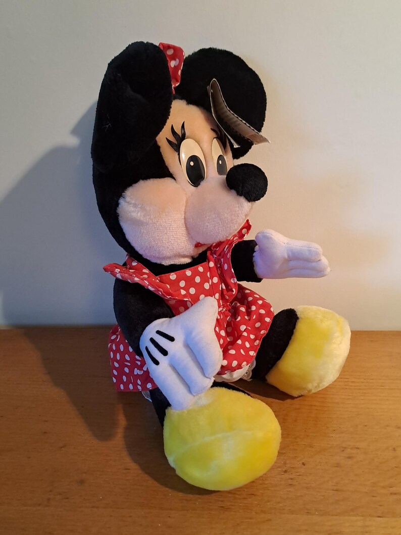 May include: A plush Minnie Mouse toy wearing a red dress with white polka dots. She has black ears with a red bow, black eyes, and yellow shoes.