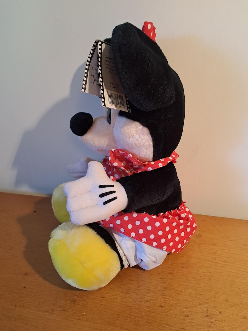 May include: A plush toy of a cartoon mouse wearing a red and white polka dot dress and a black beret. The mouse is sitting with its legs crossed and has a yellow and black tail.