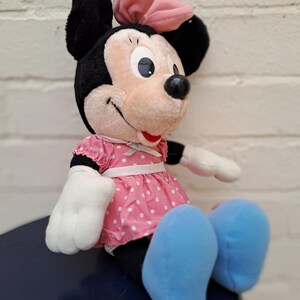 May include: A plush Minnie Mouse doll, featuring black ears, a pink bow, and a pink polka-dot dress. The doll has white gloves and blue shoes, with a smiling expression. The doll is sitting on a dark blue surface.