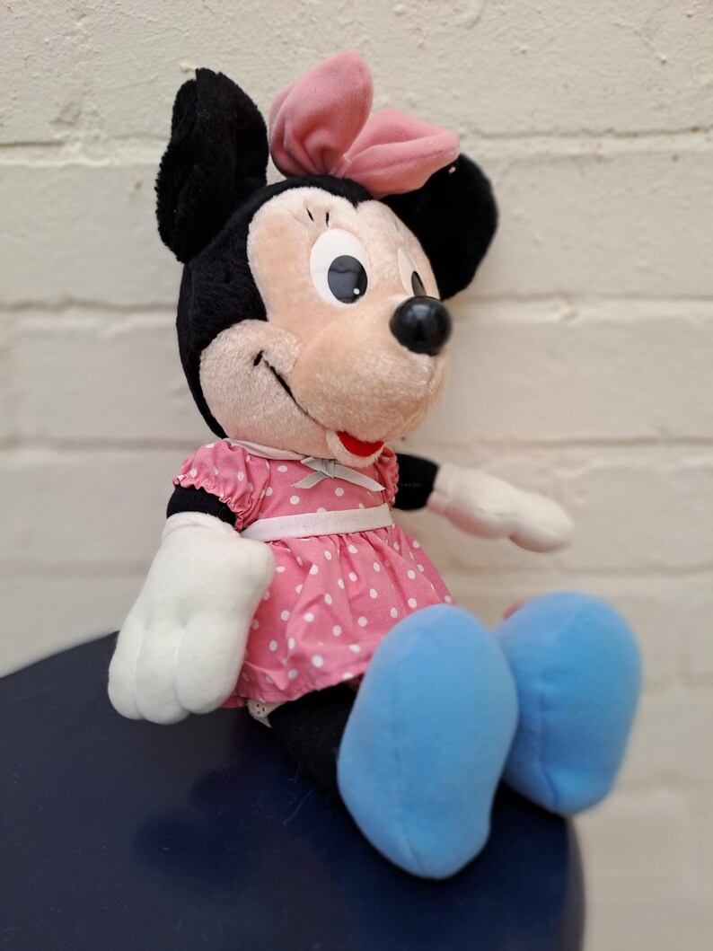 May include: A plush Minnie Mouse doll, featuring black ears, a pink bow, and a pink polka-dot dress. The doll has white gloves and blue shoes, with a smiling expression. The doll is sitting on a dark blue surface.