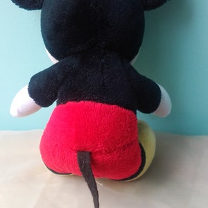 May include: A black and red plush Mickey Mouse toy with a yellow face and large black ears. The toy is sitting on a white surface.
