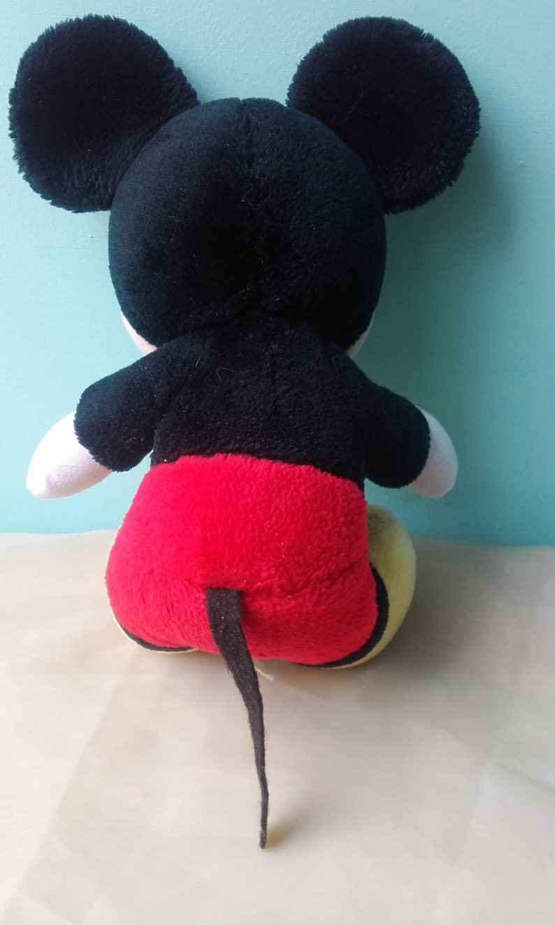 May include: A black and red plush Mickey Mouse toy with a yellow face and large black ears. The toy is sitting on a white surface.