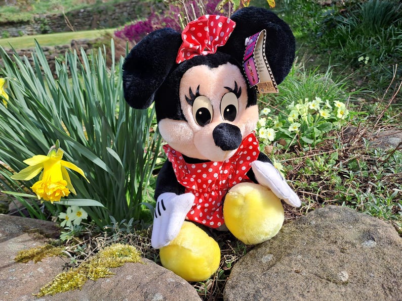 May include: A plush Minnie Mouse toy wearing a red dress with white polka dots and a red bow. She has large black eyes and a black nose. The toy is sitting on a rock with green grass and yellow flowers in the background.