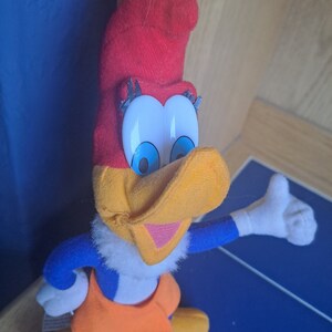 May include: A plush toy of Woody Woodpecker, a cartoon character. The toy is red, yellow, blue, and white. Woody is wearing a red hat and has a big smile.