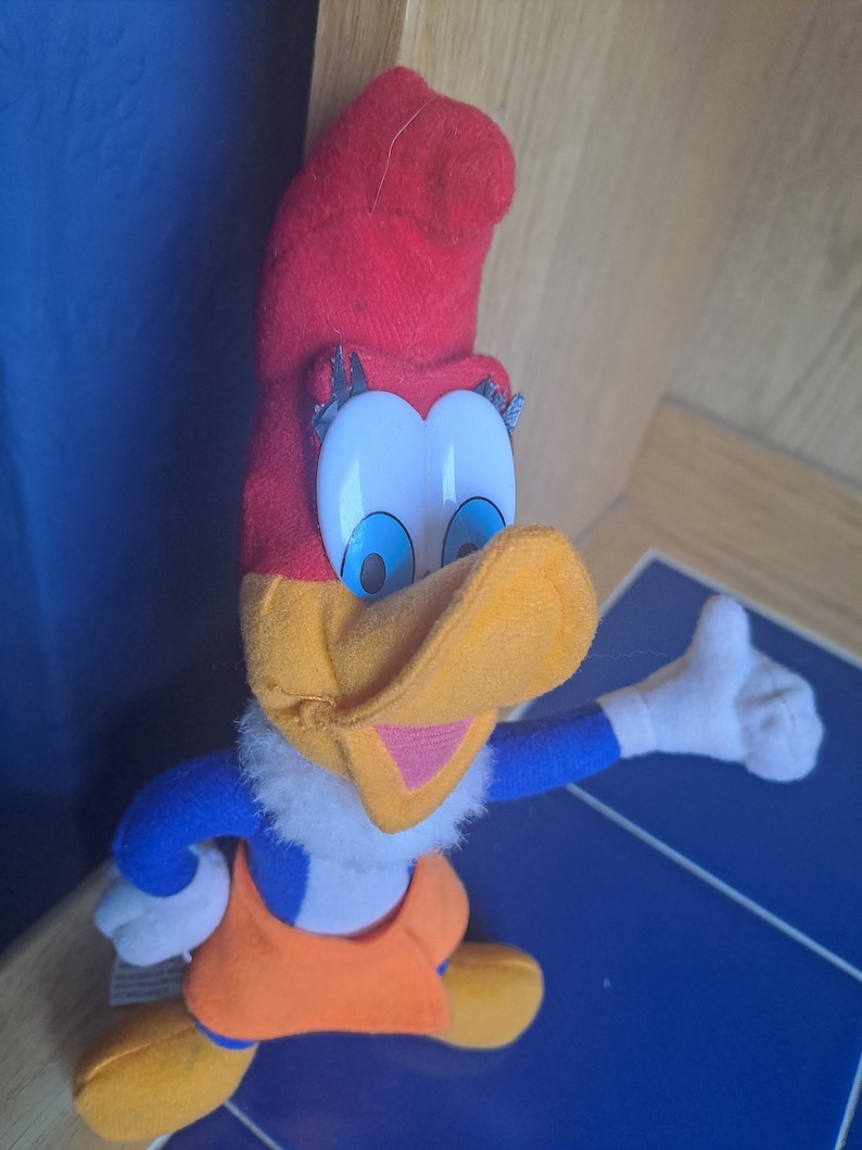 May include: A plush toy of Woody Woodpecker, a cartoon character. The toy is red, yellow, blue, and white. Woody is wearing a red hat and has a big smile.