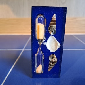 May include: A blue hourglass with seashells embedded in the resin. The hourglass is filled with orange sand and has a clear glass bulb.