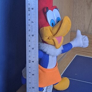 May include: A plush toy of Woody Woodpecker wearing a blue and white jumpsuit with an orange skirt. The toy is standing on a blue surface with a ruler in the background. The ruler is marked in inches.