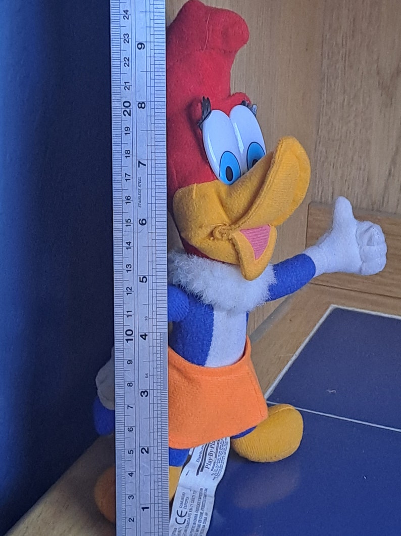 May include: A plush toy of Woody Woodpecker wearing a blue and white jumpsuit with an orange skirt. The toy is standing on a blue surface with a ruler in the background. The ruler is marked in inches.