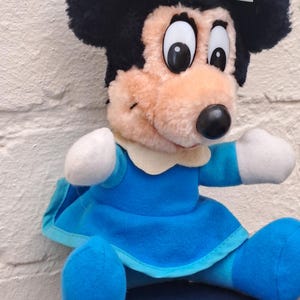 May include: A plush Minnie Mouse doll with black fur, large eyes, and a light blue bow. She wears a blue dress with white accents and blue shoes. The doll is sitting down.